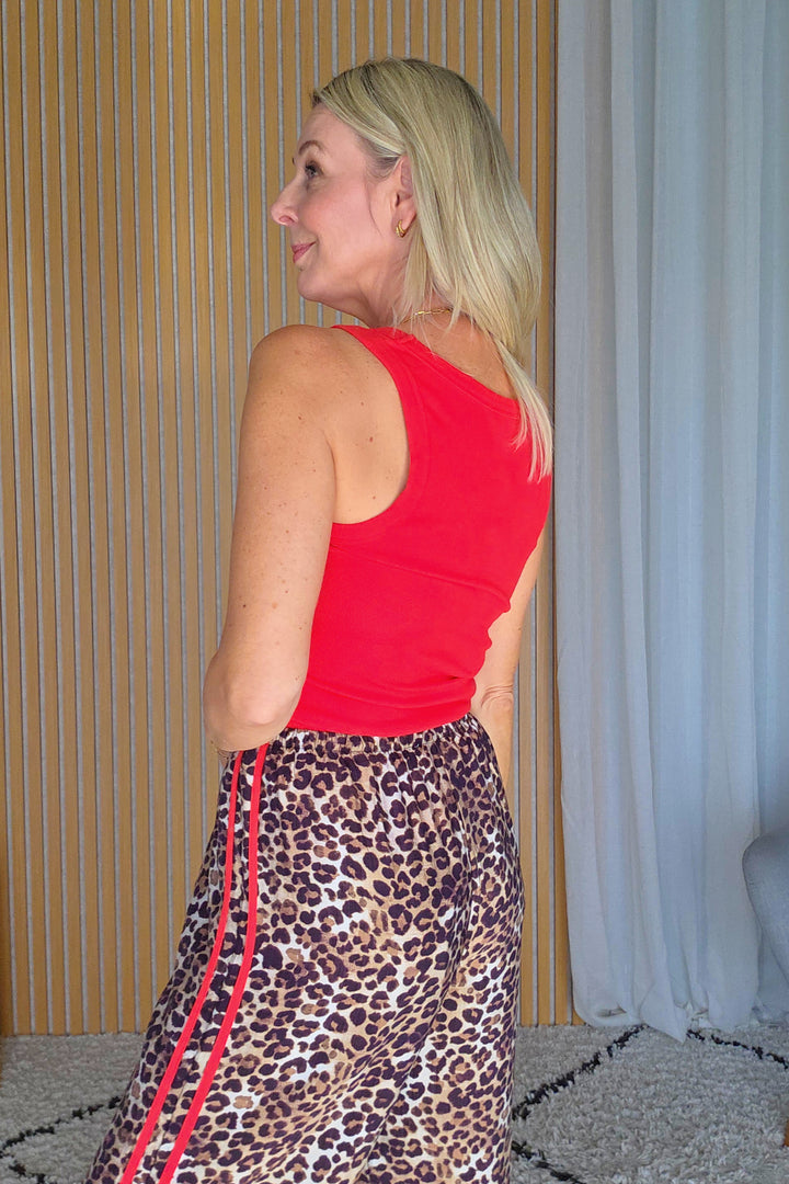 A woman wearing a bright red sleeveless top and a leopard-print skirt with red side stripes, standing indoors against a wood-paneled wall and white curtain.